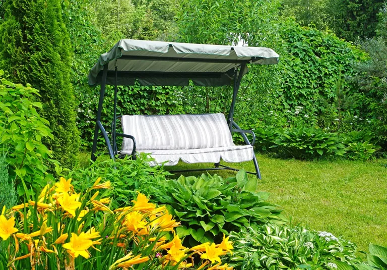 Huśtawka ogrodowa z daszkiem wśród zieleni i kwiatów w przydomowym ogrodzie